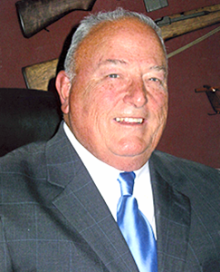 A man in a suit and tie stands confidently in front of a gun rack, showcasing a serious demeanor and professional attire.