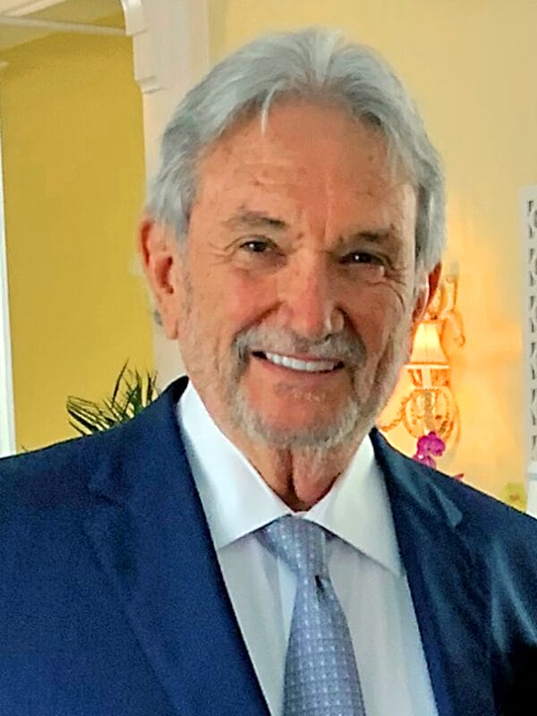 A man with gray hair wearing a blue tie, standing confidently with a neutral expression.