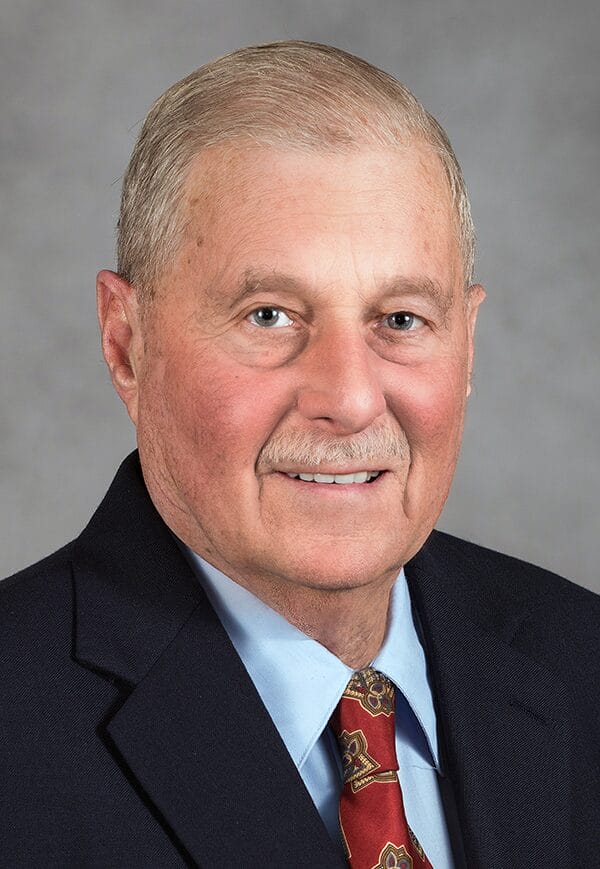 Older white male wearing a dark suit jacket, white collared shirt, and a red tie.