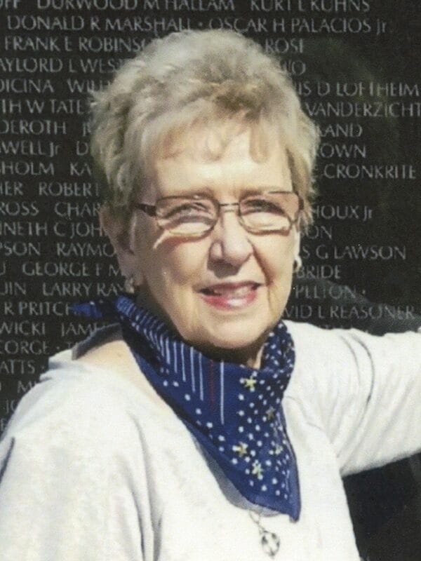 An older woman stands beside a memorial wall, reflecting on the names and tributes engraved on its surface.
