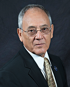 A man wearing a formal suit and tie, standing confidently with a neutral expression.