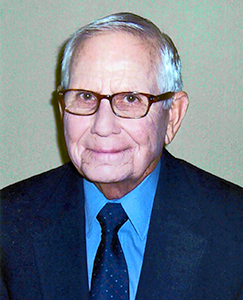An older man with glasses wearing a blue shirt, smiling and looking directly at the camera.