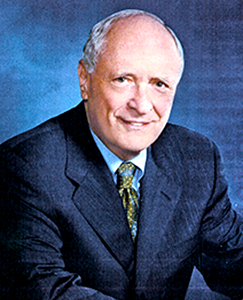 A man in a suit and tie seated at a desk, focused on his work with a laptop and documents in front of him.