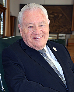 A man in a formal suit is seated comfortably in a chair, looking relaxed and composed.
