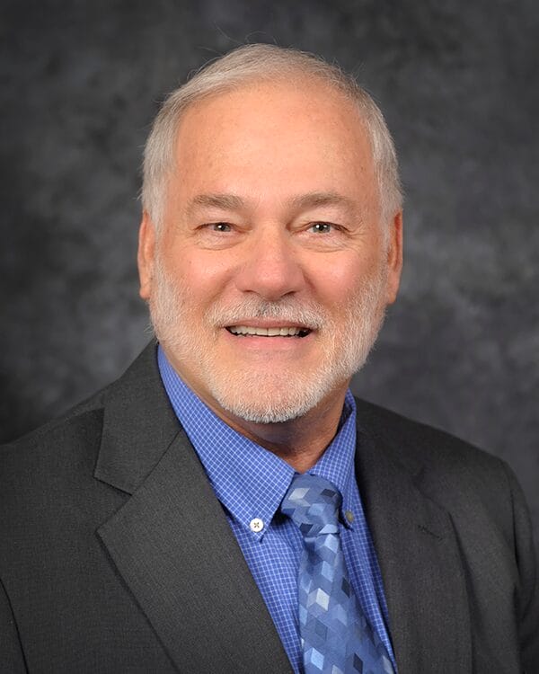 A man with a gray beard wearing a blue shirt and tie, looking confidently at the camera.