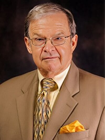 A man wearing a suit and tie, glasses, and a bright yellow tie, standing confidently with a neutral expression.