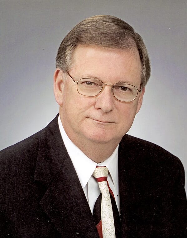 Older white male wearing glasses, a suit jacket, white collared shirt, and a tie.