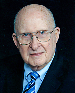 A man in a suit and tie seated at a desk, focused on his work with a laptop and documents in front of him.