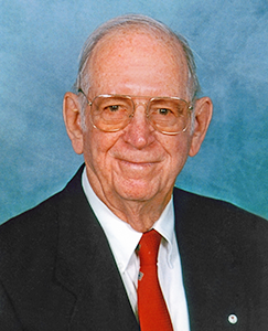 An older man wearing a suit and a red tie, standing confidently with a neutral expression.