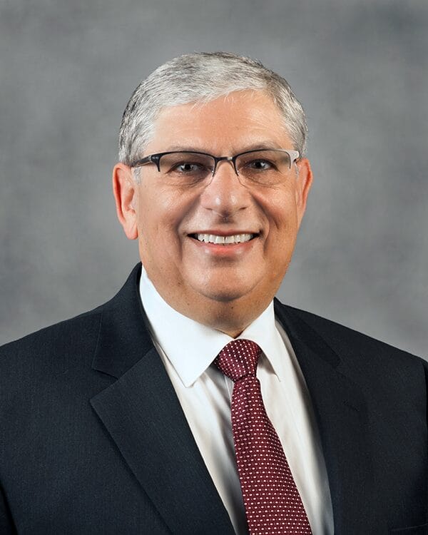 Older male with light brown skin. He wears black glasses, a suit and tie.