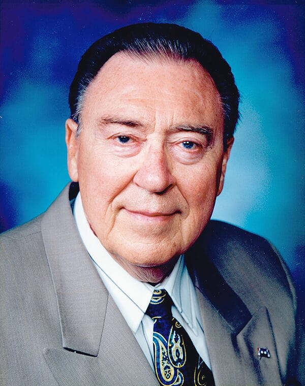 Older white male with slicked back dark hair. He is wearing a grey suit and tie.