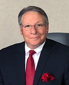 An older man with glasses wearing a blue shirt, smiling and looking directly at the camera.