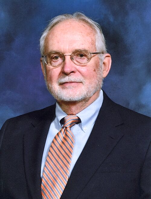 A man wearing glasses and a tie, dressed in a formal suit, stands confidently with a neutral expression.