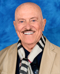 A bald man wearing a mustache and a scarf, looking directly at the camera with a neutral expression.