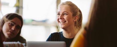 blonde student smiling while sitting at a table with other students, with a laptop in front of her