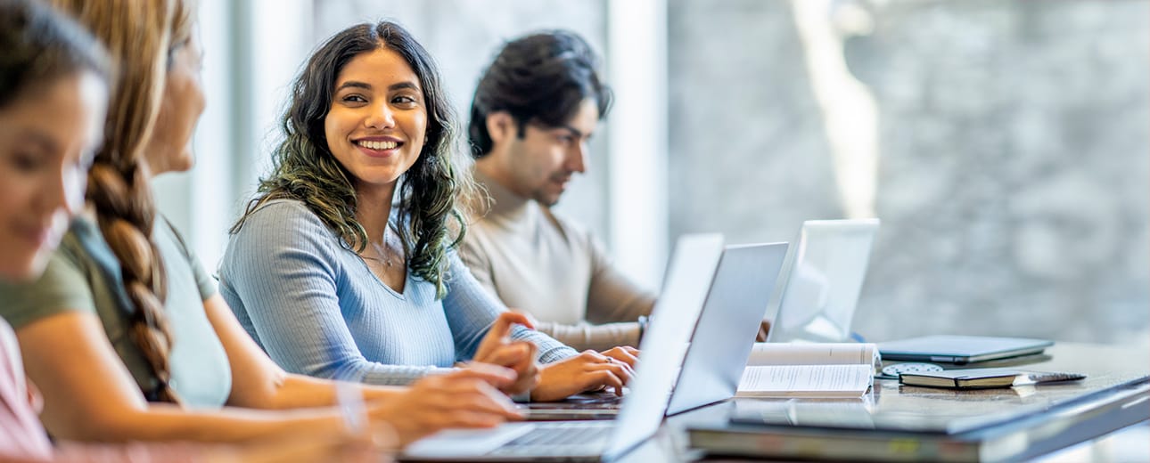 A group of college students work on laptops at a long table, while a young woman in a blue sweater smiles at a classmate.