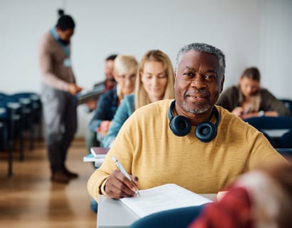 older student smiling while sitting in a classroom with headphones around his neck and pen in hand