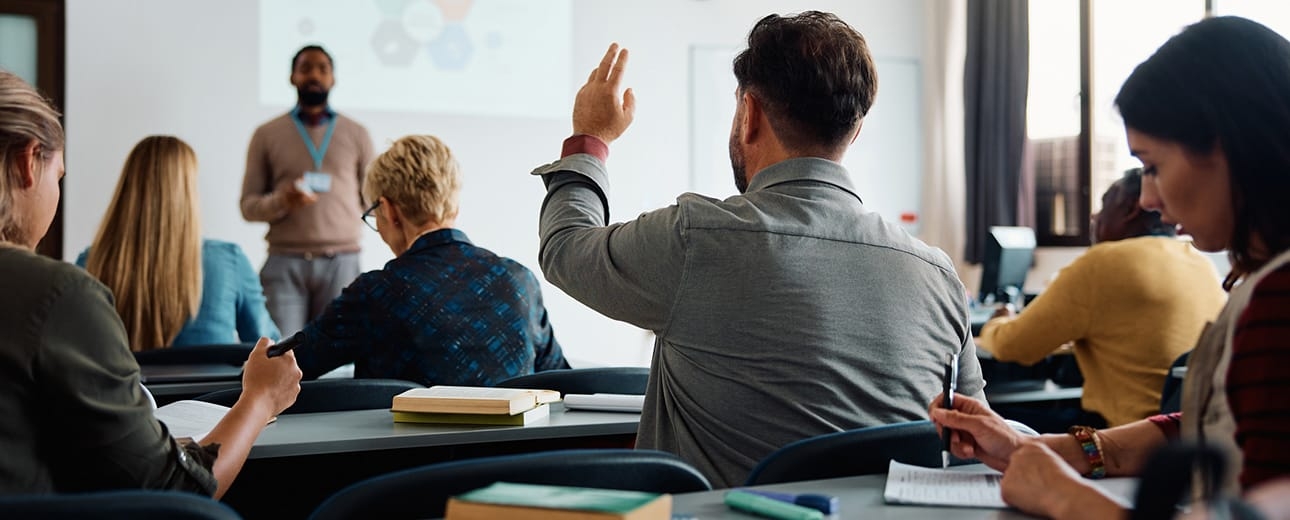 A classroom full of adult students listens to a standing instructor at the front of the room, while one man raises his hand to ask a question.