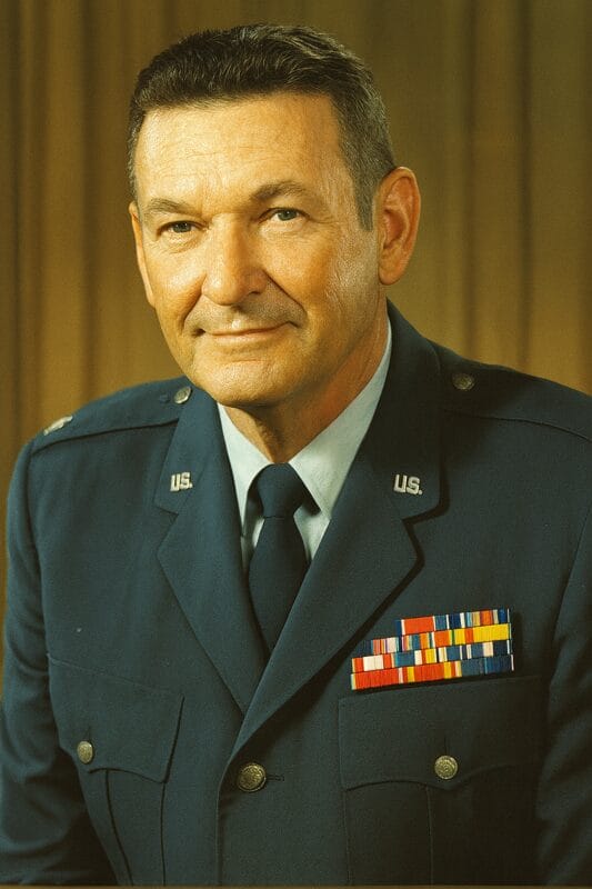 Older man in U.S. Air Force dress uniform with rank insignia and colorful service ribbons, smiling slightly in front of a neutral curtain backdrop.