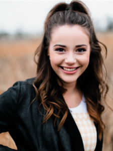 Young woman with long wavy brown hair in a half-up style, smiling and wearing a dark jacket over a yellow gingham dress, standing in a field with tall grass.