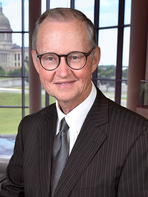 Smiling older man in glasses and a dark pinstripe suit with silver tie, posed in front of large windows overlooking a capitol building and landscaped grounds.