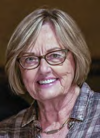 Smiling older woman with short blonde hair and glasses, wearing a plaid shirt and a necklace, posed indoors with a dark background.