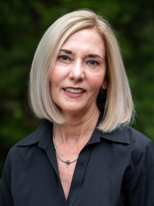 Smiling woman with straight blonde hair in a chin-length bob, wearing a black collared shirt and a delicate necklace, posed outdoors with a blurred green background.