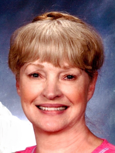 Smiling older woman with light blonde hair styled in bangs and a braided crown, wearing a bright pink top, posed in front of a soft blue studio backdrop.