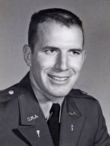Young man in military uniform with short hair, smiling in a black-and-white studio portrait.