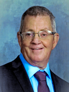 Older man with short gray hair and glasses, smiling in a dark suit with a light blue shirt and maroon tie, posed in front of a blue textured backdrop.