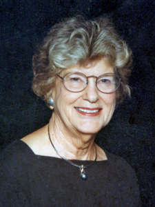 Older woman with short wavy gray hair and glasses, smiling and wearing a black top with a pearl pendant necklace, posed against a dark backdrop.
