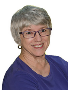Smiling older woman with short gray hair and glasses, wearing a purple top and small hoop earrings, posed against a white background.
