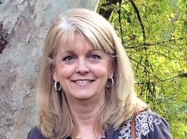 Smiling woman with long blonde hair, wearing hoop earrings and a dark jacket with embroidered detailing, standing outdoors in front of a tree and leafy background.