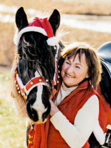 Smiling woman in a red vest hugging a black-and-white horse wearing a Santa hat and festive halter, outdoors in a sunny field.