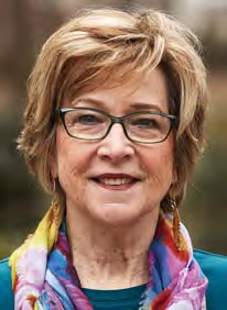 Smiling older woman with short light brown hair and glasses, wearing a colorful scarf over a teal top, posed outdoors with a blurred natural background.