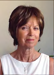 Older woman with short dark brown hair and side-swept bangs, wearing a white sleeveless top and a cross necklace, posed against a neutral indoor background.