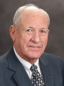 Older man with short white hair, wearing a blue suit jacket, white shirt, and patterned tie, posed against a soft gray studio backdrop.