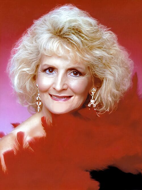 Smiling older woman with voluminous blonde hair, sparkly dangling earrings, and a red feather boa, posed against a red and pink studio backdrop.