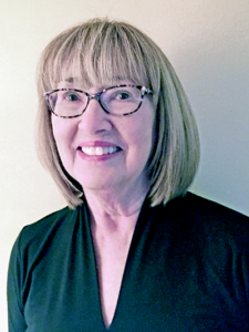 Smiling woman with straight blonde hair in a bob with bangs, wearing glasses and a black V-neck top, posed against a light-colored wall.