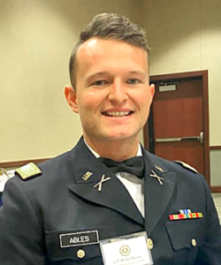Smiling man in formal military dress uniform with name tag reading “Ables” and multiple service ribbons, standing indoors at an event with a badge around his neck.