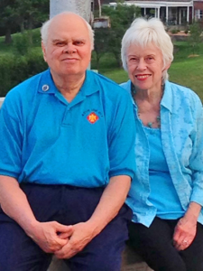 Elderly man and woman sitting close together outdoors, both smiling. The man wears a bright blue polo shirt and the woman wears a matching light blue blouse. A grassy field and a large house are visible in the background.