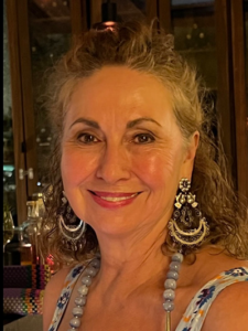 Older woman with short curly gray hair and glasses, smiling and wearing a sleeveless black top with a silver embellished neckline, seated in a banquet setting.