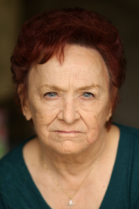 Older woman with short auburn hair and a serious expression, wearing a teal top and a delicate necklace, posed against a dark blurred background.