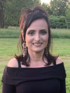 Smiling woman with dark shoulder-length hair, wearing dangling earrings and an off-the-shoulder black top, standing in a grassy field with trees in the background.