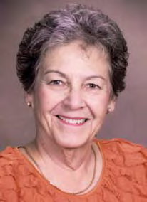 Smiling older woman with short curly gray hair, wearing a textured orange top and small stud earrings, posed against a soft mauve backdrop.