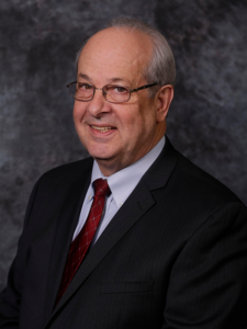 Older man with glasses and thinning gray hair, smiling in a dark suit with a red patterned tie, posed against a mottled gray studio backdrop.