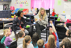 Elementary kids surrounding two college girls reading a book.