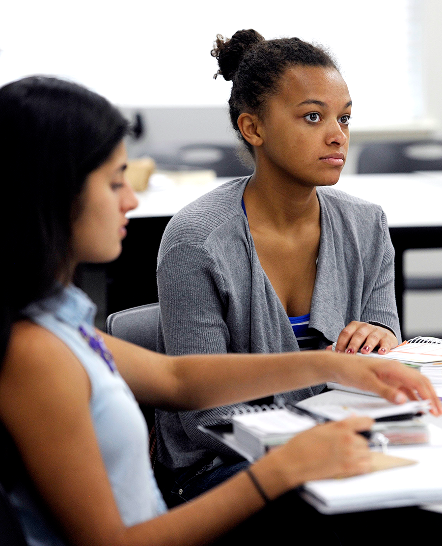 Two girls in class listening to a lecture and taking notes.
