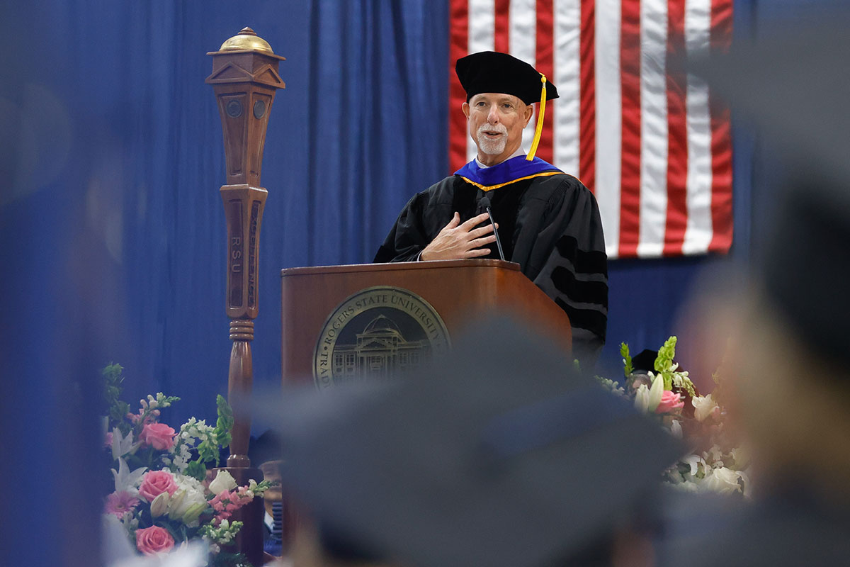 man in cap and gown speaking at podium