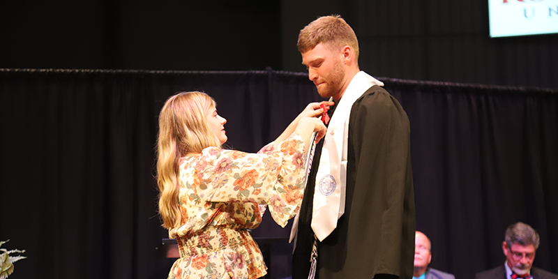 Woman pinning ribbon to male student in graduation gear